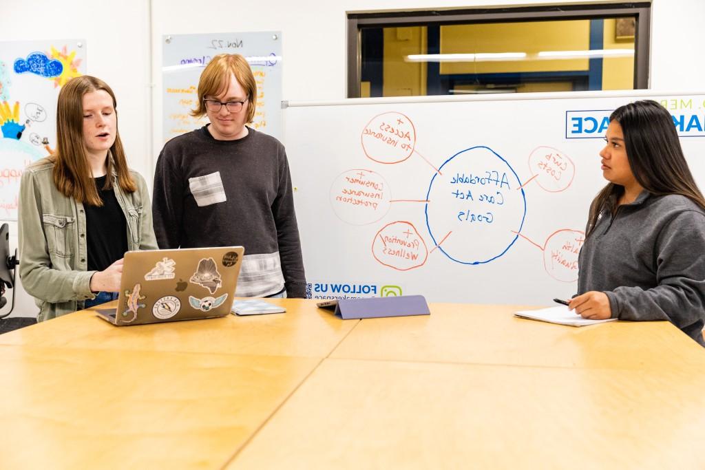 Three U N E students stand at a table in front of a whiteboard