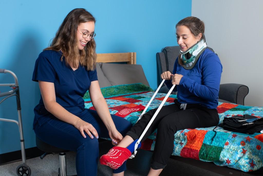 An Occupational Therapy student wearing blue scrubs assists a patient wearing a neck brace with a foot sling